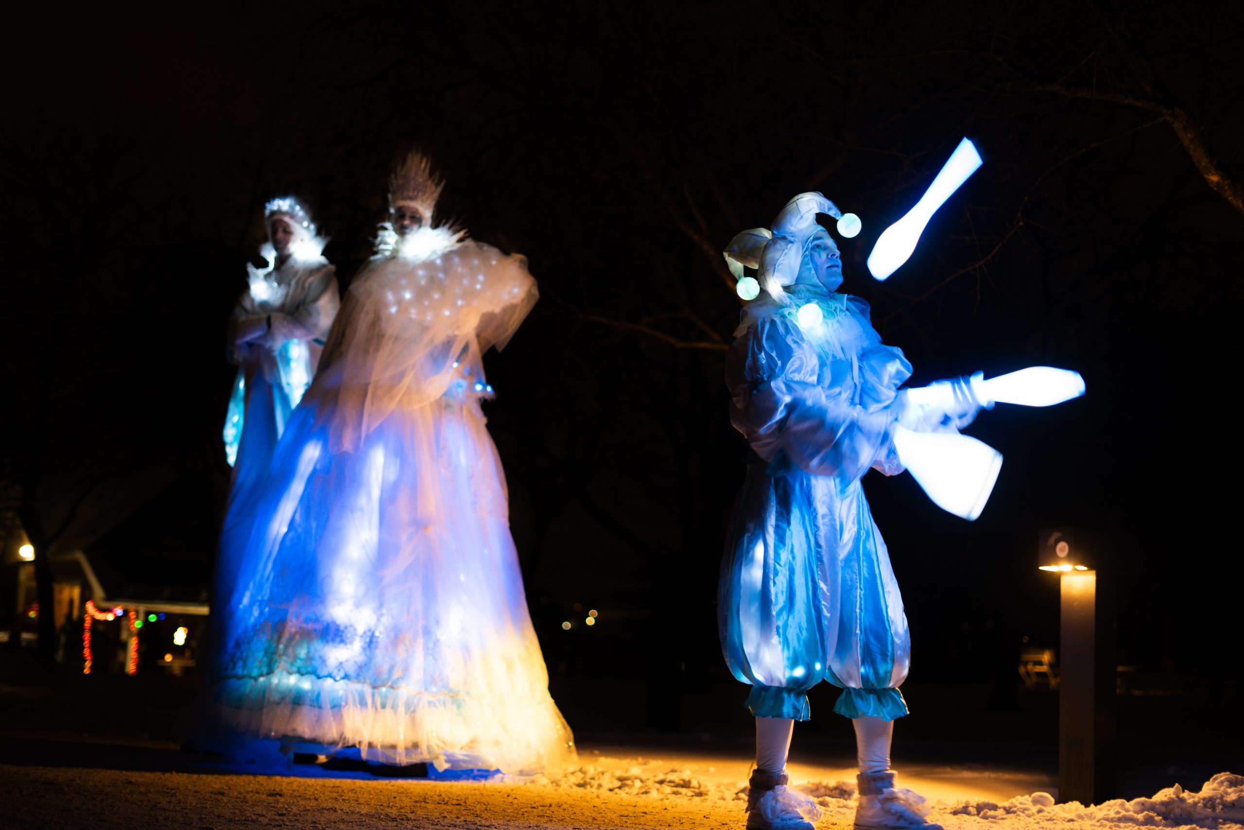 Siniseen pukuun pukeutunut narri heittelee hohtavia keiloja. Taustalla seisoo kaksi korkeaa hahmoa, joilla näyttää olevan jäiset kruunut päässään. Maassa on lunta.